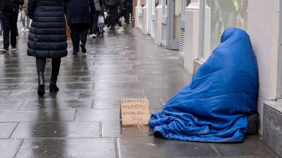 A homeless person sits on the pavement hidden under a blue sleeping bag while people pass by on a wet Oxford Street in London. Beside them is a cardboard sign saying 'I am very hungry,  God bless you'