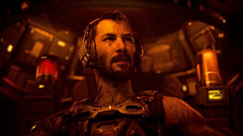 A battle-scarred man sits inside the futuristic cockpit of a robot craft, illuminated in a dim orange light emanating from the controls. He wears a communications headset and a metal harness.