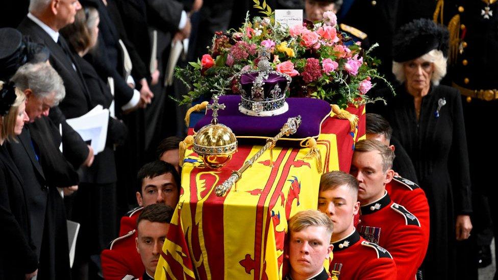 Men in military red uniform carrying the Queen's coffin, draped in a flag, holding flowers, the crown, an orb and a sceptre.