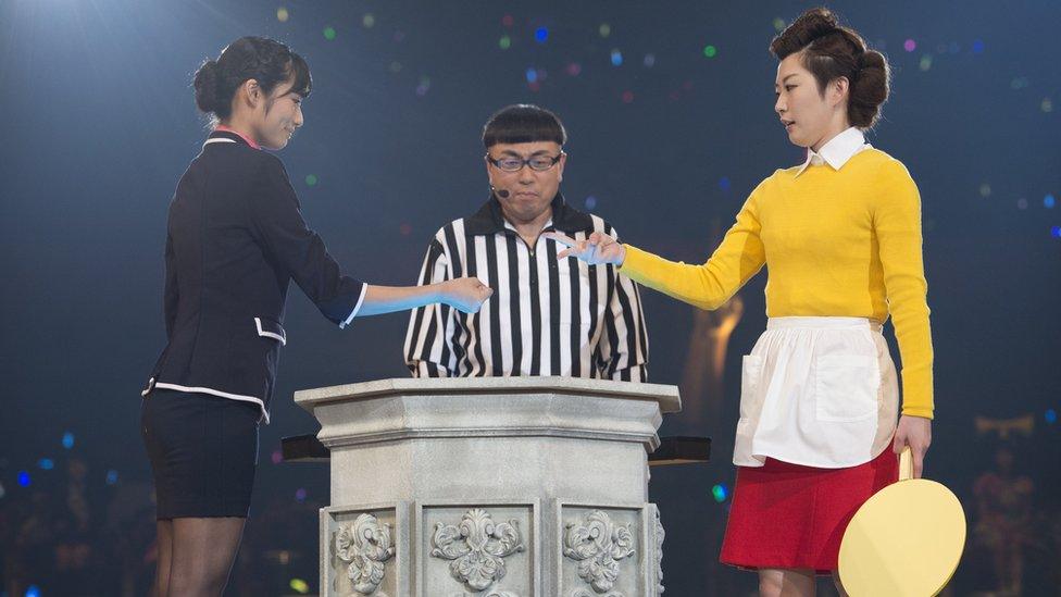 Two band members compete during the rock-paper-scissors tournament in 2015, with an umpire looking on. The woman on the right has just thrown "scissors", losing to "rock" on the left.