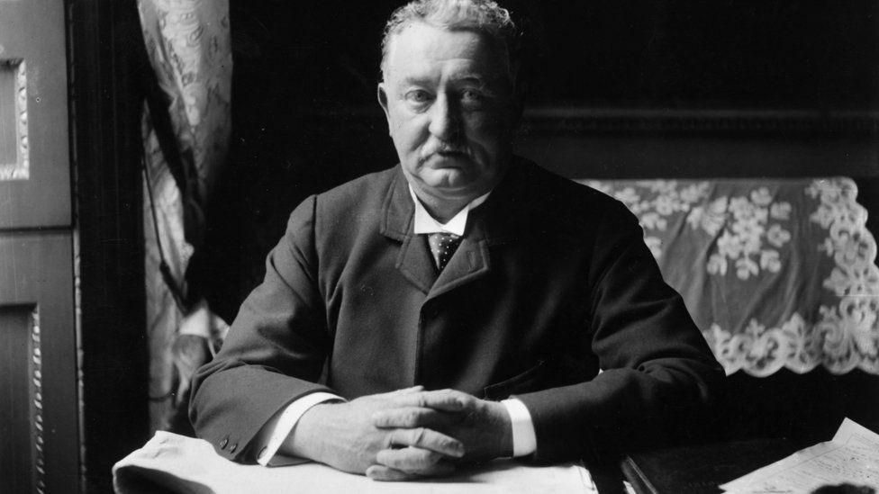 A black and white photo of Cecil Rhodes sat at a desk, his hands clasped together. He wears a Victorian-era suit.