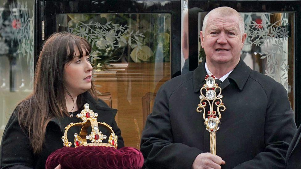 A woman holding the jewelled crown on a red velvet cushion, and a man holding the sceptre, standing in front of the hearse