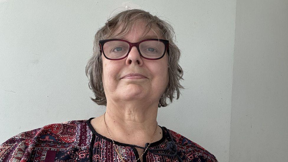 Jackie Moore has hair above her shoulders and is wearing glasses and a pink and purple top and is looking straight at the camera