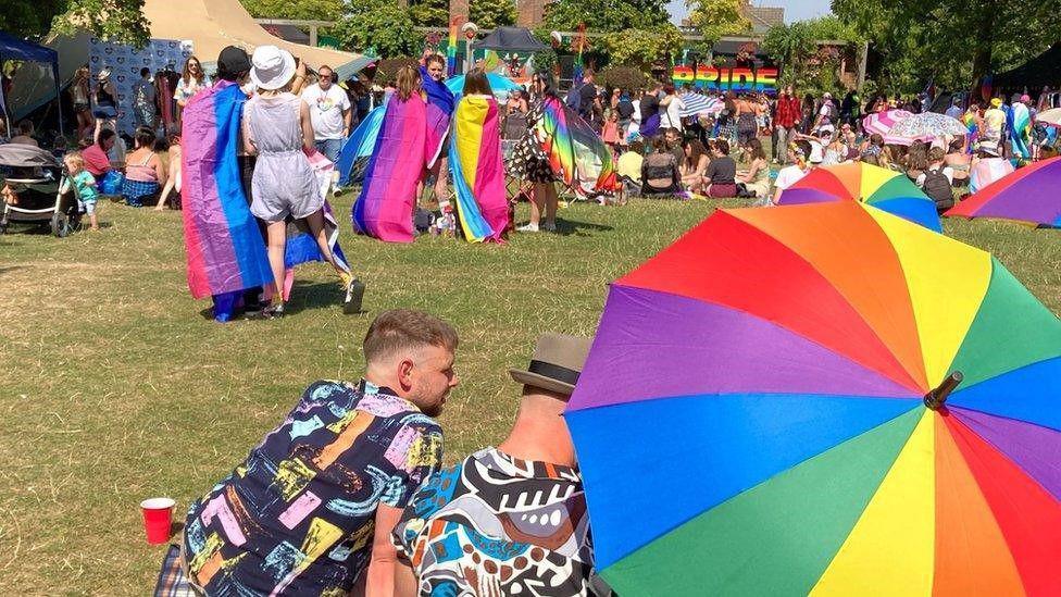 Two men wearing patterned shirts are sitting on the grass in front of a rainbow-coloured umbrella. One is wearing a green trilby hat. In front of them is a crowd of people in colourful outfits, some with Pride flags, drinking and taking in the sunshine. There are some people sitting on blankets and some children in prams. In the background is a sign which says "PRIDE" in rainbow lettering.