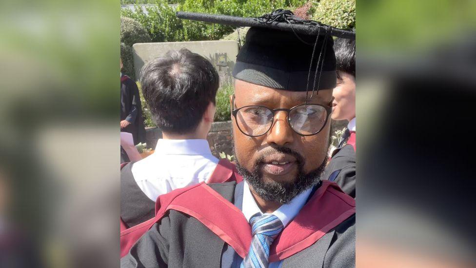 A selfie of Abdullahi Abdi wearing his black graduation robes with a maroon hood and a blue striped tie. He is wearing a black mortarboard with a hanging tassel and black framed glasses. He is looking at the camera and behind him there are other students in their graduation robes outside on a sunny day. 