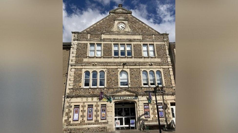 The Gateway Theatre in Seaton pictured on a sunny but cloudy day. The front of the theatre appears to be three to four storeys with a large clock front on the fourth storey. The entrance has a large sign reading THE GATEWAY above the doorway. 