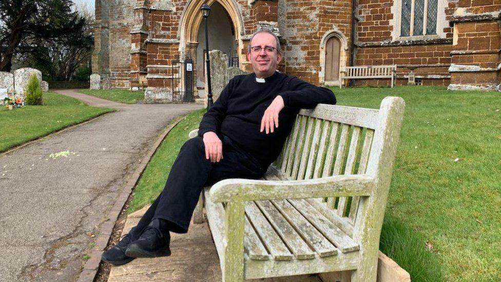 Richard Coles in clerical clothing with white collar sitting outside a church