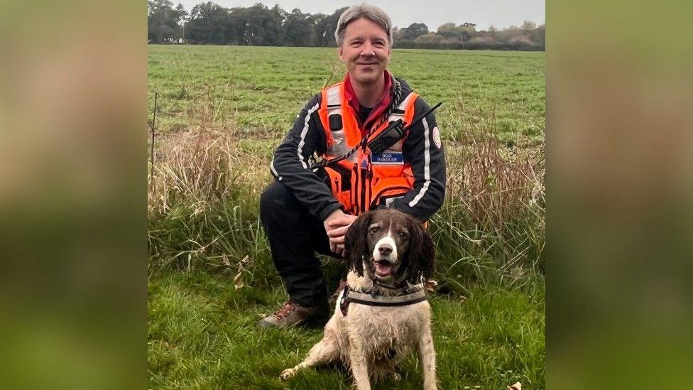 Millie, a brown and white Springer Spaniel, sitting in front of her handler Darren Yeates in a field. Darren is wearing a bright orange high viz jacket with walkie talkie attached.