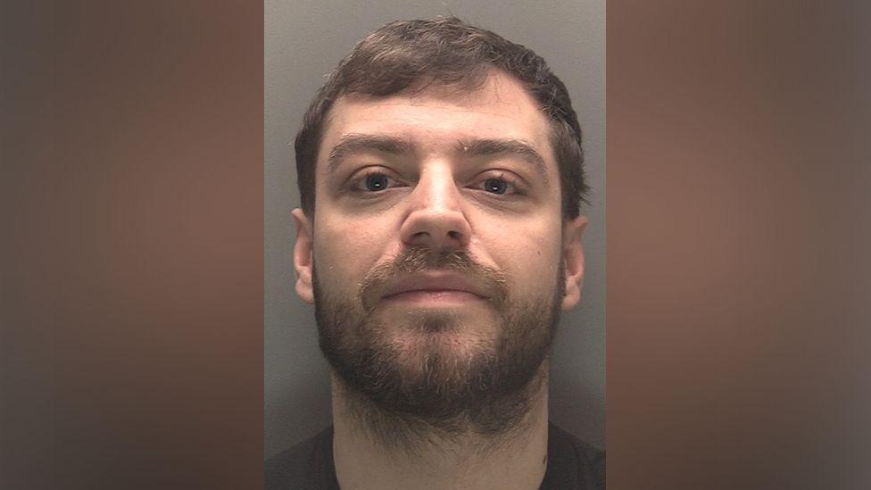 A police mugshot of Lee Harrison, 27, who has brown hair and a brown beard and smirks at the camera.