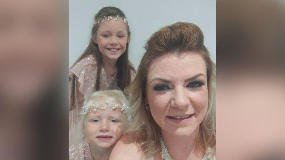 Vicky Parker smiling at the camera with her two young girls beside her. The two girls are wearing princess dress-up clothing. 