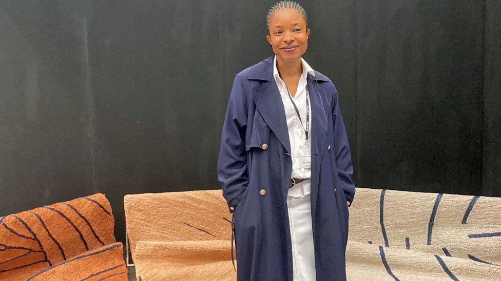 Kelechi Ejikeme in a long navy coat standing in front of jute rugs - mainly brown and beige with navy lined designs on them - at the Decorex show in London