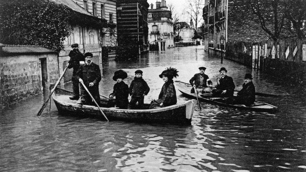 people in heavy old-fashioned outfits and hats on boats in a narrow city street