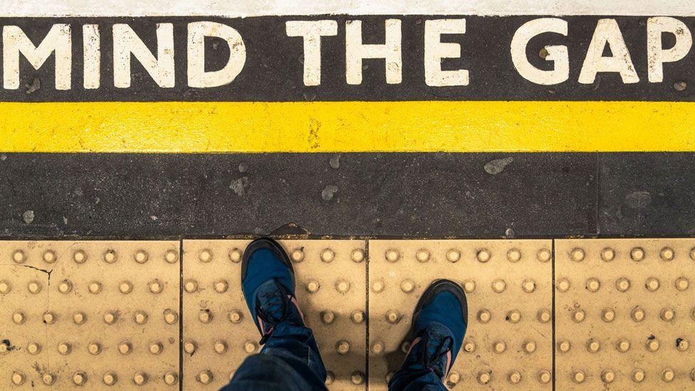 Mind the Gap sign shot taken vertically so you can see the sign on the platform and someone wearing trainers standing behind the yellow line on the bobbled paved section. Above the yellow line there is a Mind the Gap sign painted in white bold capital letters.