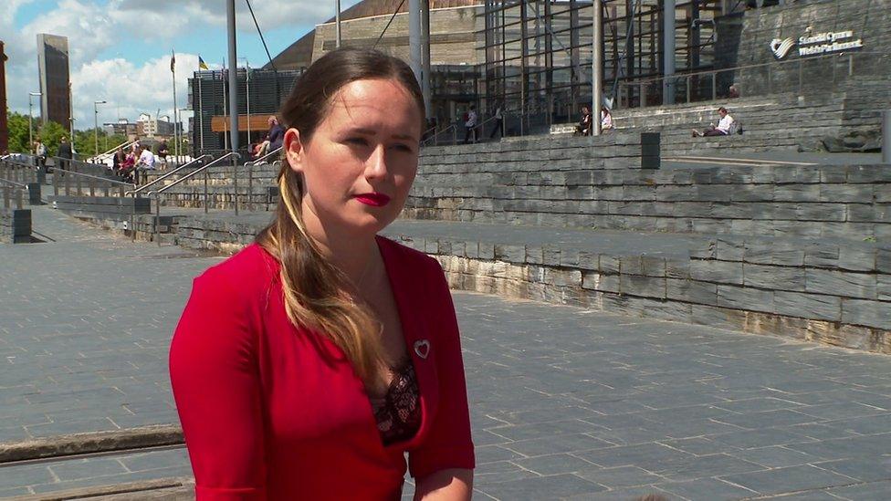 A woman in a red dress with long, dark hair sits outside the Senedd in Cardiff Bay.