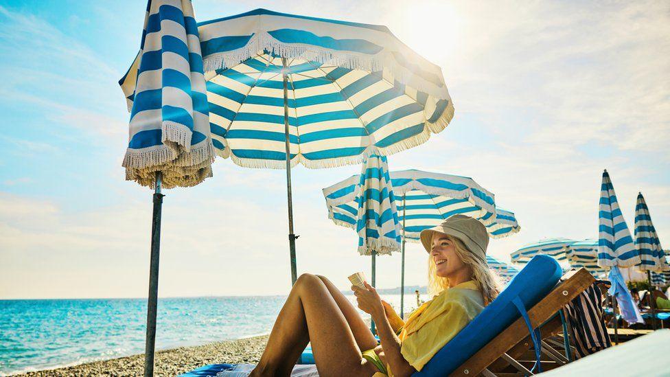 A woman sits on a sun lounger under a sun umbrella on a beach.