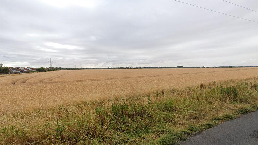 Google street view of part of the proposed site showing a field of crops with housing in the distance and electricity pylons overhead