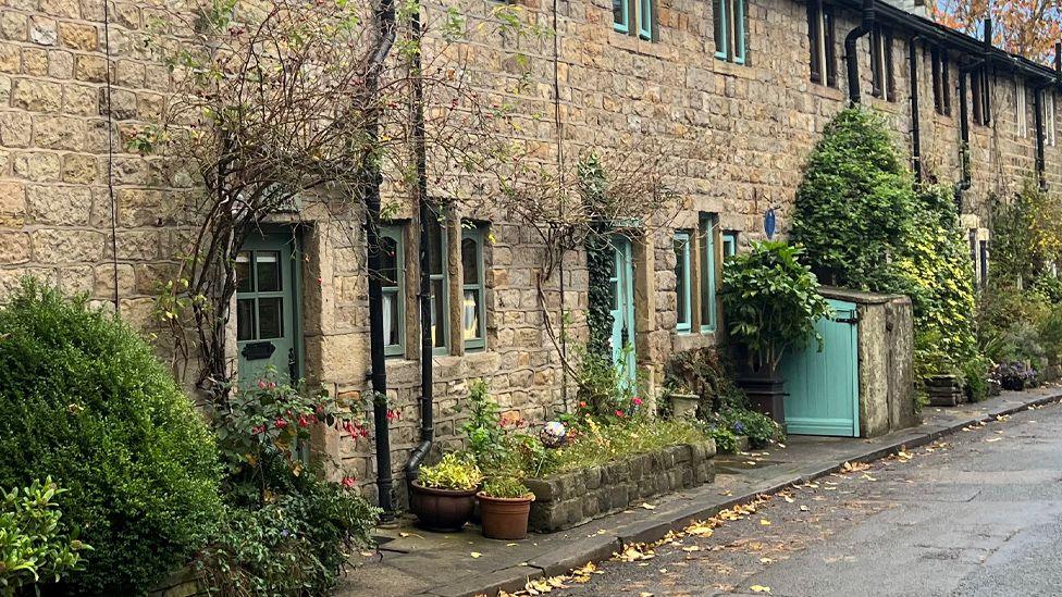 A pretty row of stone cottages, with green doors and windows and lots of plants and flowers along the front