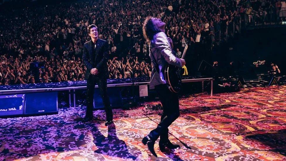 Brandon Flowers and guitarist Dave Keuning look up at a screen on stage showing the end of the match