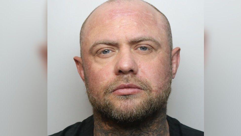 A mugshot of Reece Marshall, who has a bald head, facial hair and tattoos on his neck