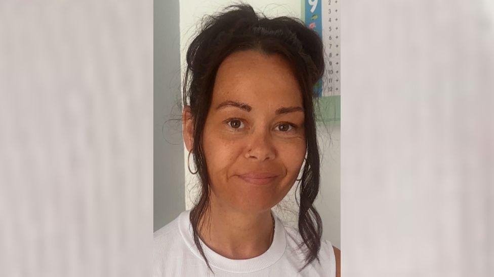Rebeca Proctor has black hair in a bob and brown eyes. She is wearing a white top and is smiling at the camera. 