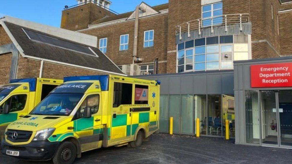 Two ambulances parked up at the entrance to a hospital building with large glass doors to the right and a emergency department sign above.