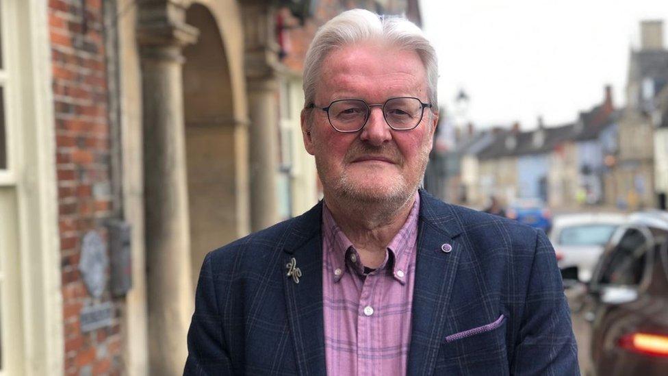 A grey haired man with grey spectacles in a pink checked shirt and blue checked suit jacket looking straight at the camera