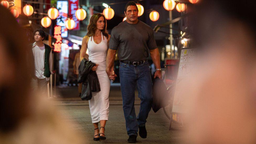 Dwayne Johnson and Emily Blunt in The Smashine Machine. She is wearing a long white dress and Johnson is wearing a dark grey T-shirt and blue jeans.