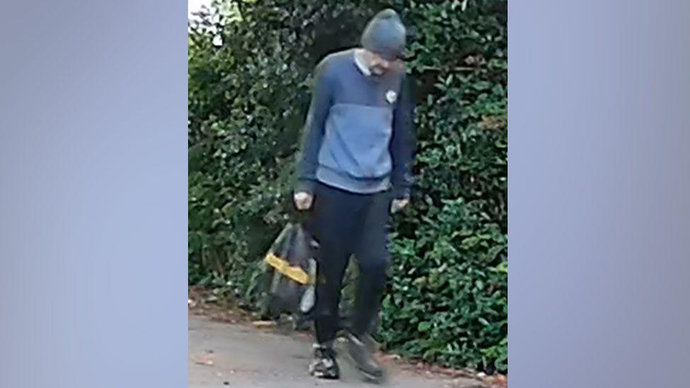 A CCTV image of an unidentified man walking in the village of Brantham. He wears a black hat, blue jumper, black trousers and black shoes.