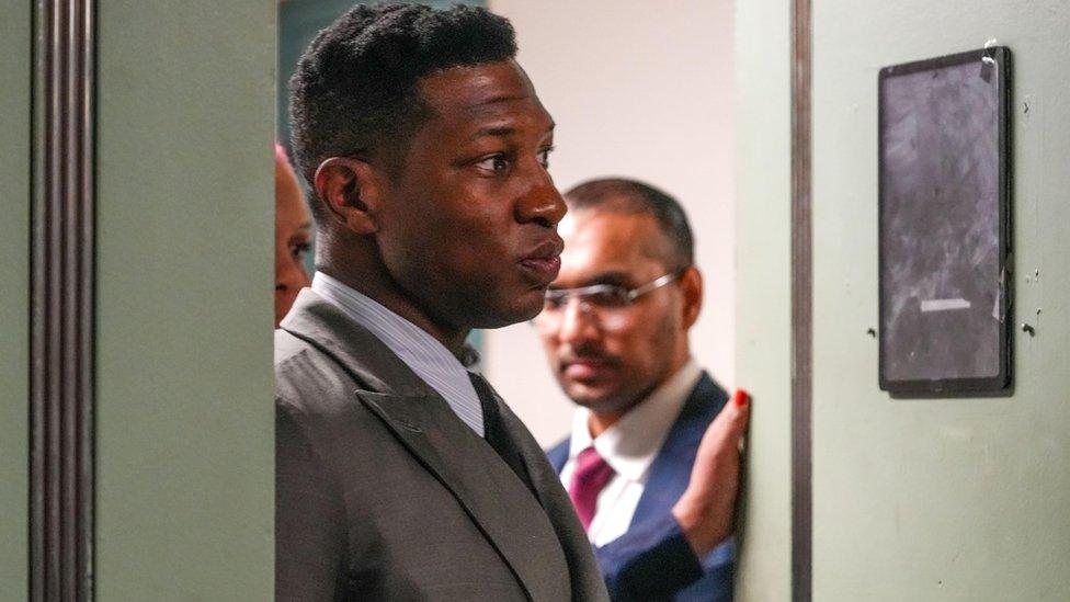 Jonathan Majors in grey suit with striped shirt.