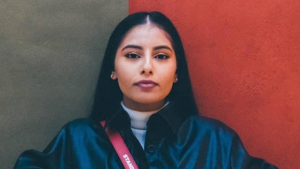 Jana Ally stood against a backdrop that is half black and half red on either side of her head. She is looking at the camera and her dark hair is worn down over the back of her shoulders. She is wearing a white roll neck jumper and a buttoned up black jacket over it. There is also a red bag strap visible over her right shoulder.