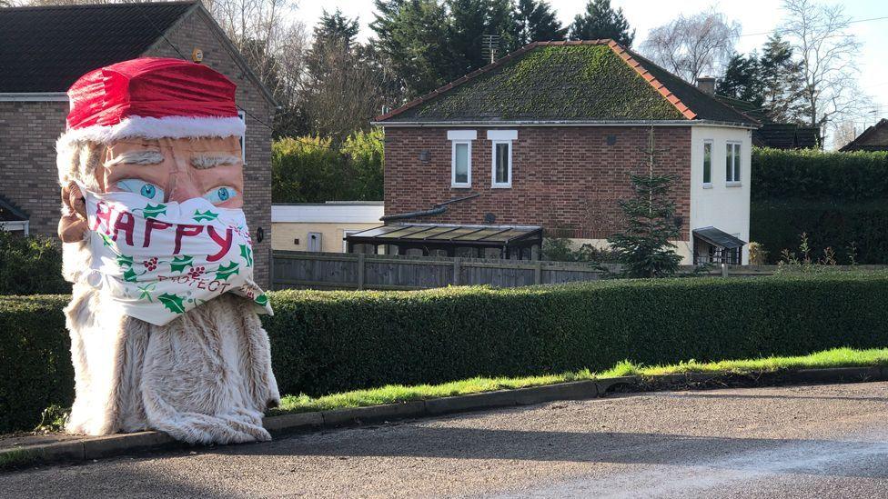 A sculpture of a large Santa face wearing a mask. The Santa figure has a red hat on and a fake fur beard. The mask is painted with the words "Happy Xmas" and "protect each other"
