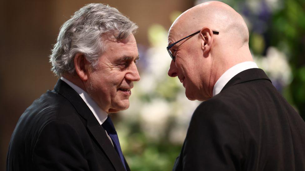 Gordon Brown, on the left of the shot with grey hair, and John Swinney, bald with glasses, talk while standing close to each other 