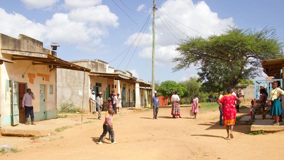 The main dirt road through the centre of Mukuku village, where some shops can be seen and residents milling around