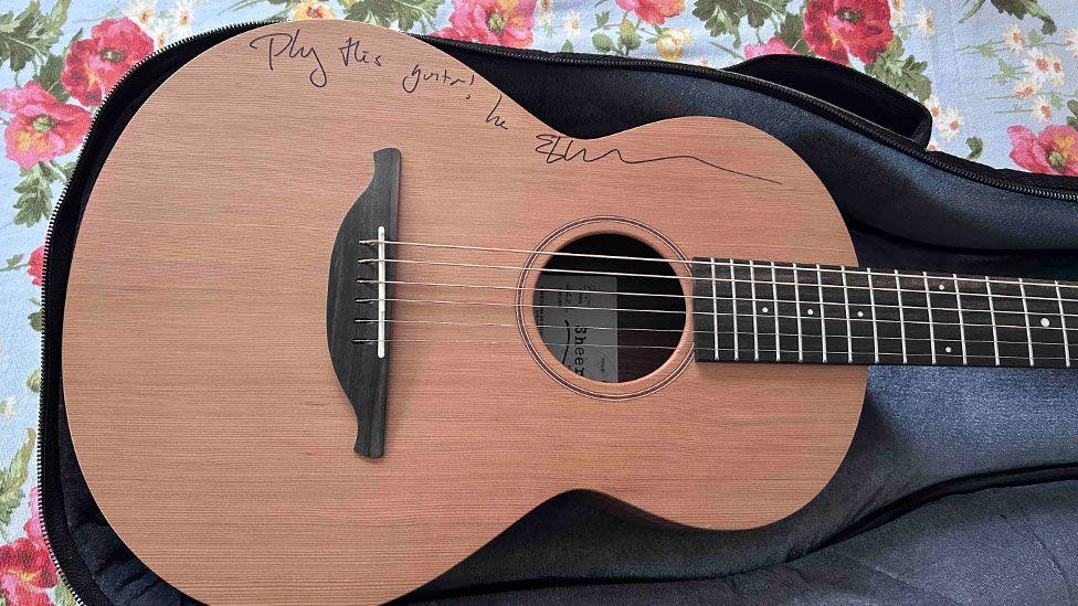 An acoustic guitar which has a light wood front and dark wood fretboard. Along its top edge is written play the guitar and it is signed by Ed Sheeran. It is resting on a black case, which is lying on flowery fabric. 