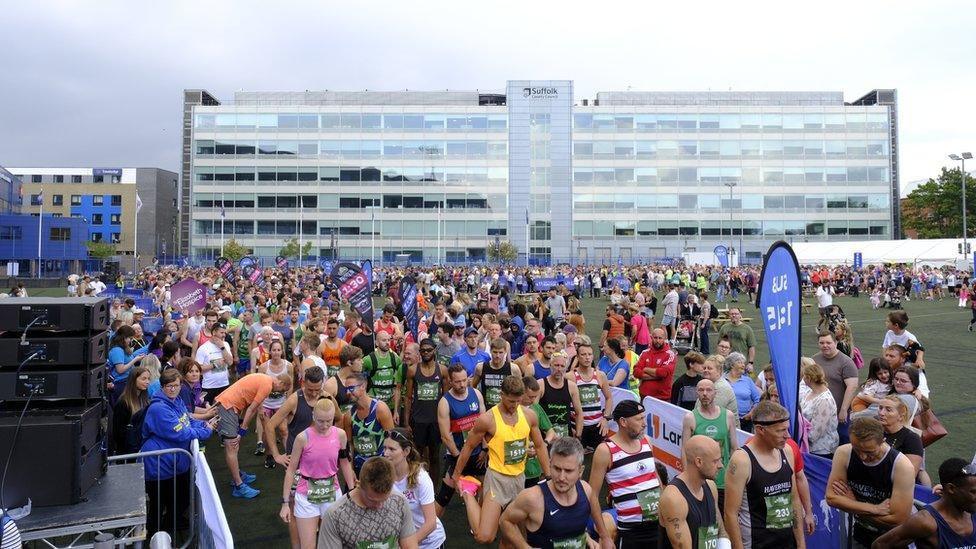Hundreds if not thousands of runners gathered together ahead of the start of the Ipswich Half Marathon. A large glass building is in the background. 