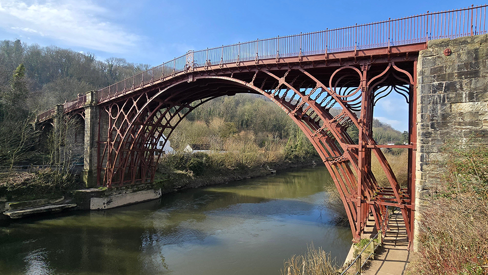 A red, arched metal bridge with decorative metalwork and metal railings at the top over a green river