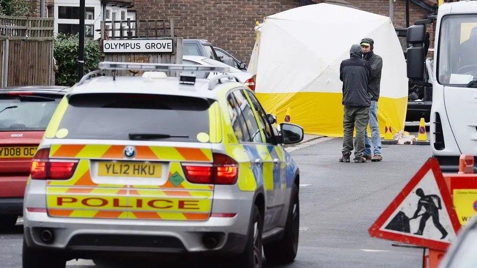 Police officers stand near a yellow and white forensic tent on a residential street in Olympus Grove, Wood Green, with a marked police car in the foreground.