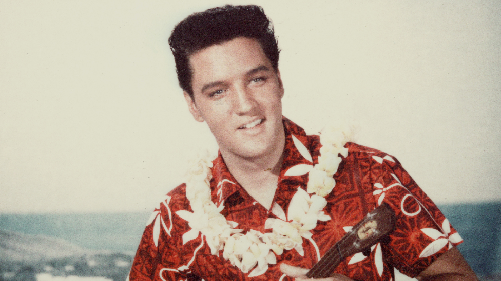 American rock n' roll singer Elvis Presley plays a ukelele, wearing a Hawaiian shirt and lei, in a still from the film 'Blue Hawaii'.