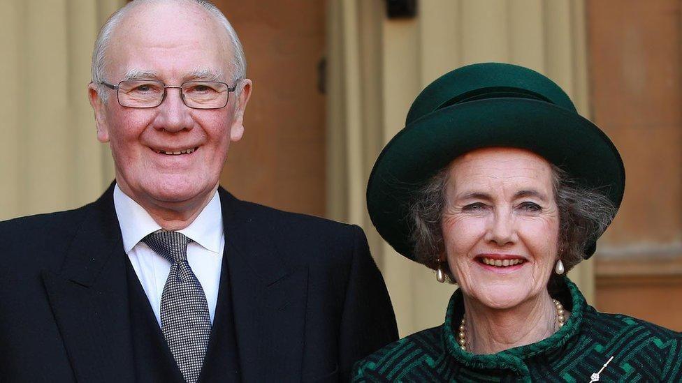 Sir Menzies is wearing a dark suit, white shirt, and tie. He is standing next to his wife Elspeth who is dressed in a green outfit with a matching hat, pearl necklace and pearl earrings.