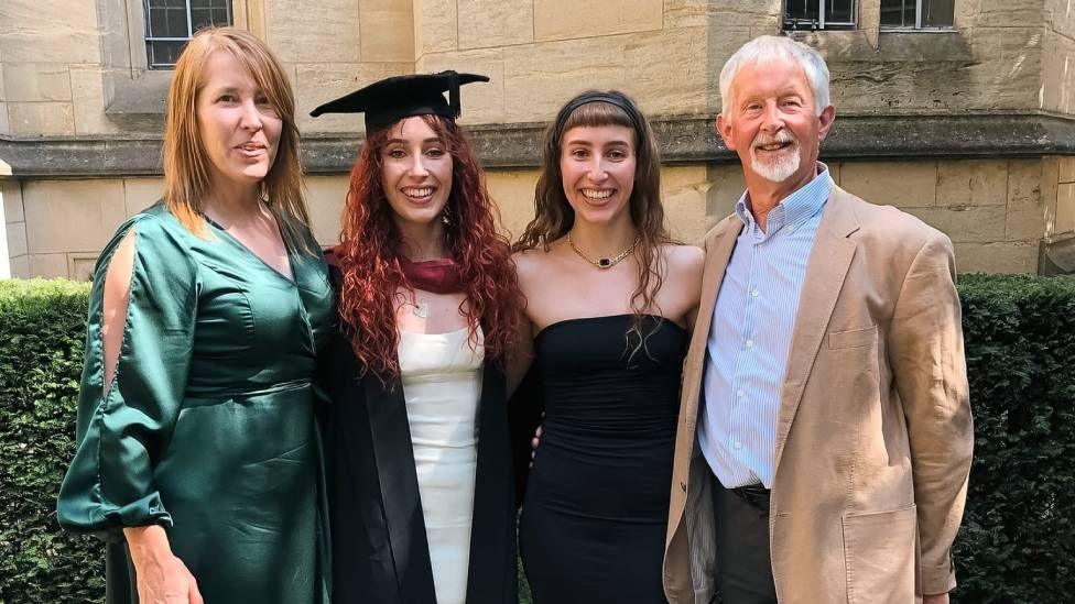 Four people lined up and smiling. On the left is a woman in a green dress, then a woman in a graduating gown, a woman in a black dress and a man in a cream coloured jacket