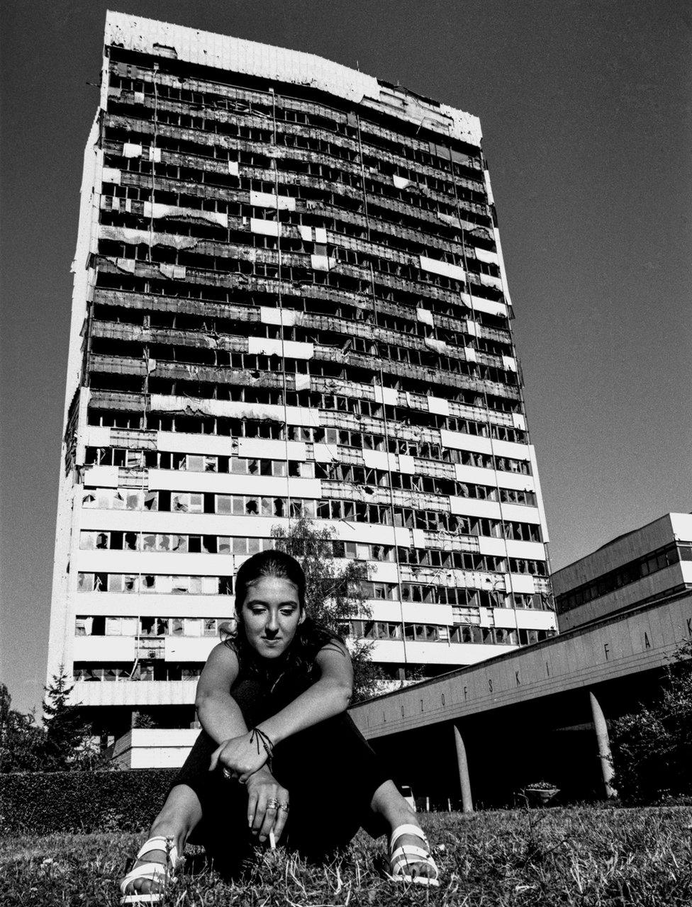 A young woman sits on the ground with a war-torn tall building behind her