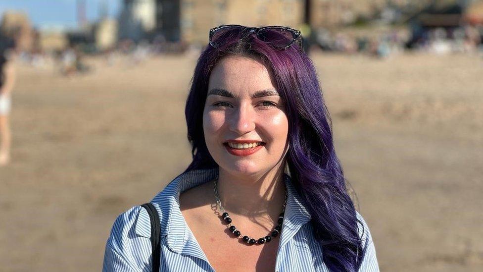 Amy Lee Fraoli stood on a beach. She is wearing a blue and white striped shirt and has her sunglasses on the top of her head