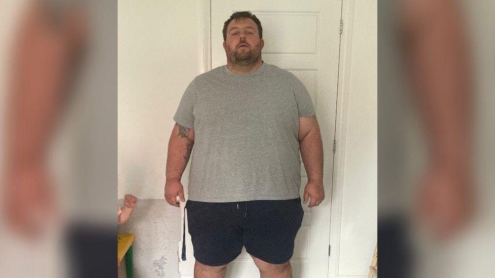 James Baker - a 38st man with brown hair and facial hair - before losing half his weight. He is wearing a grey T-shirt and black shorts.