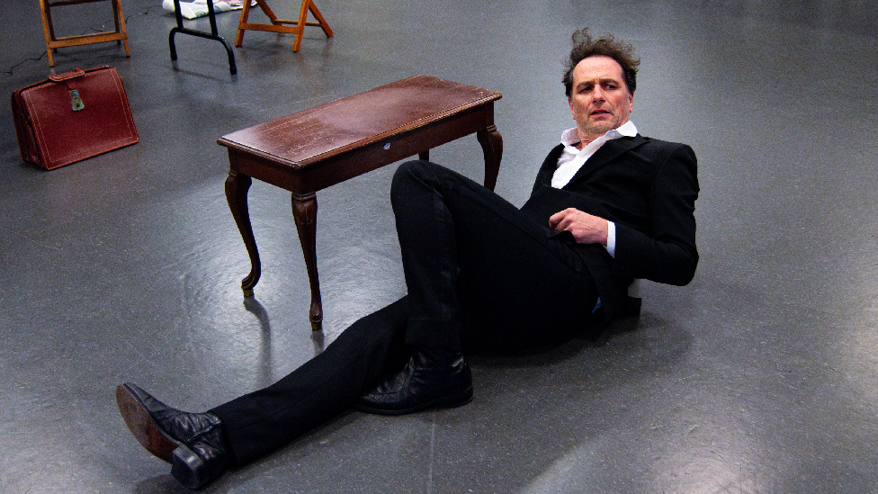 Matthew Rhys lying on the floor next to a vintaged brown wood table. He is wearing a black suit with a white shirt. His left leg is laying flat, while his right leg is upright in a 90 degrees position. He has a serious facial expression.