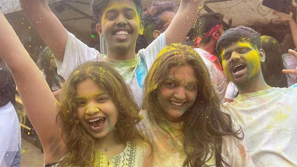 Group of four friends - two women at the front and two men at the back, celebrating the festival of Holi. There is yellow coloured powder on their faces and white shirts, and in their hair. They are shouting with their arms raised.