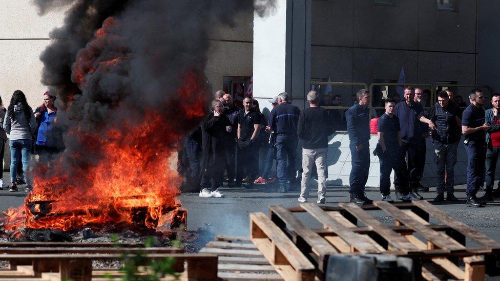 A wooden pallet burns outside the prison as guards watch on