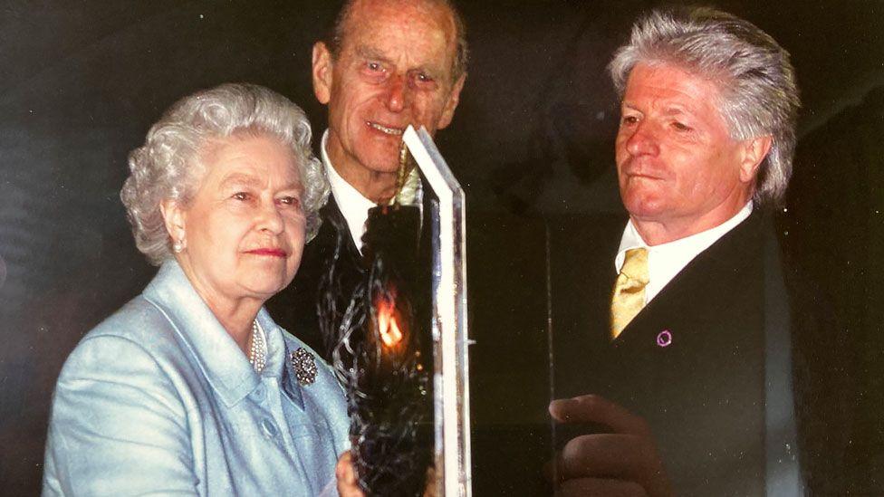 A grainy photograph of Queen Elizabeth, Prince Philip and Bruno Peek at the National Golden Jubilee Beacon, the Mall, London. She is on the left wearing pale blue, Prince Philip is in the middle and Mr Peek on the left. Both men are in dark suits, white shirts and wearing ties. In front of them is a metal stand and the Queen is holding a lit beacon