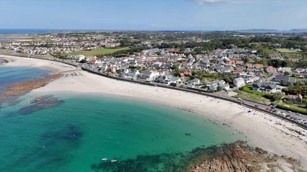A long beach with rocks, sea and sand with a breakwater and houses and fields behind.