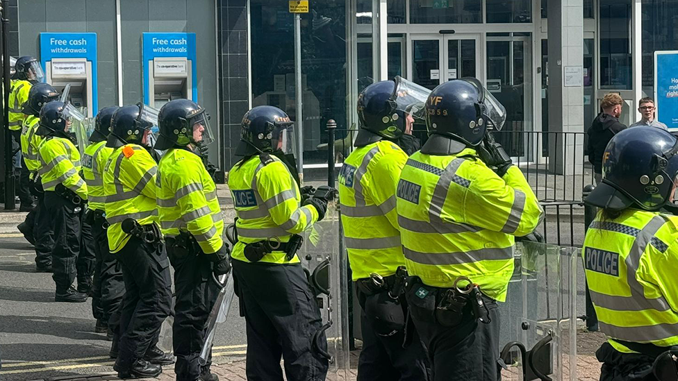 Police officers with riot shields and protective helmets standing in a line separating protesters