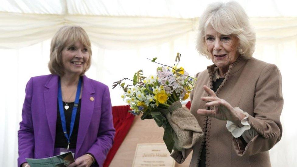 The Queen holds a bouquet of flowers, in front of the plaque and a staff member